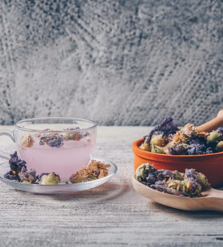 Purple colored water in a cup with dried flower tea in a bowl and spoon side view on a white wooden and gray background free space for your text